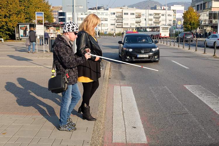 Nevidiaca žena stojí s asistentkou pred priechodom pre chodcov a zdvihnutím bielej palice signalizuje svoj úmysel prejsť cez cestu.