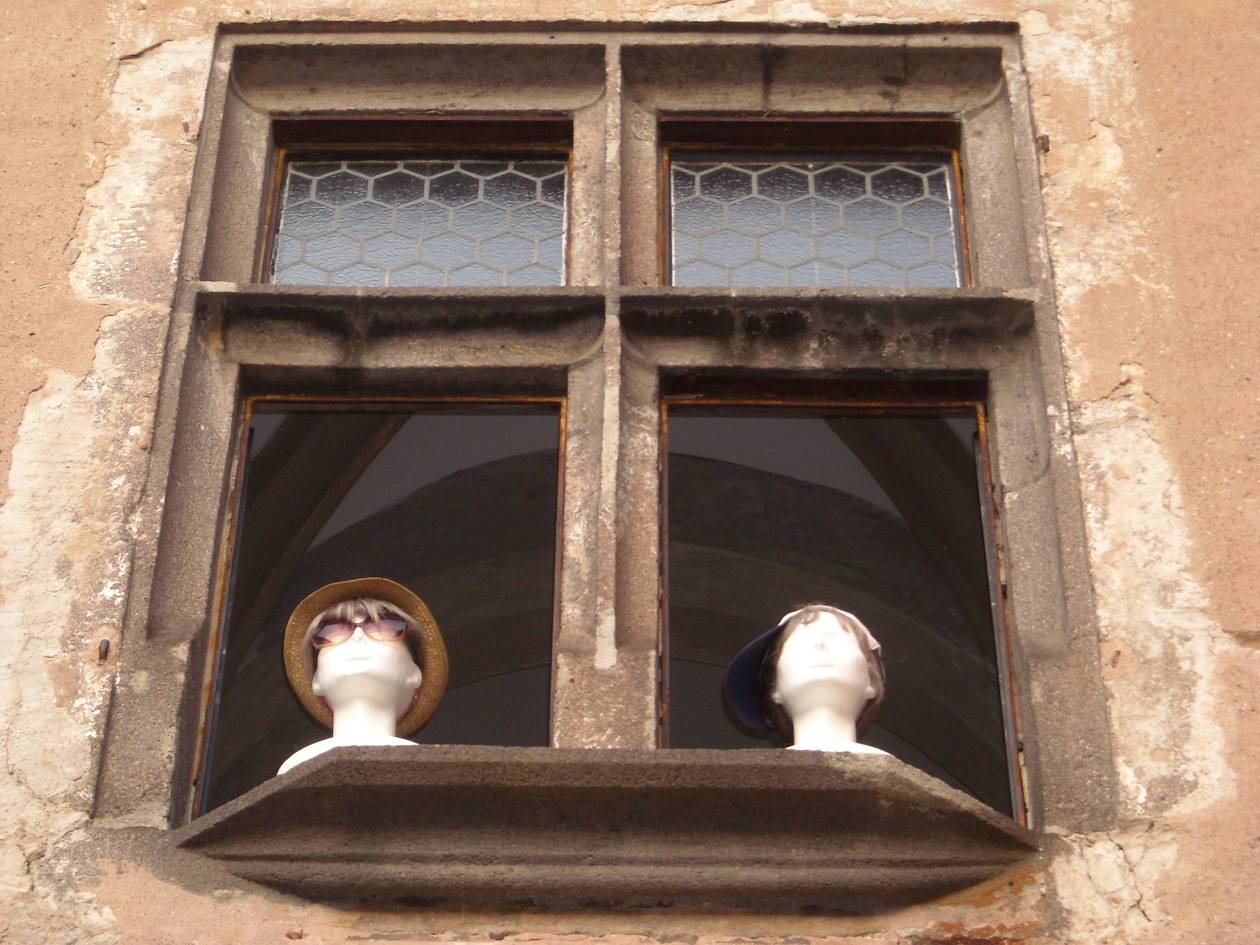 pohľad zdola na okno historickej budovy, na ktorom sú umiestnené dve plastové hlavy s klobúkom a šiltovkou