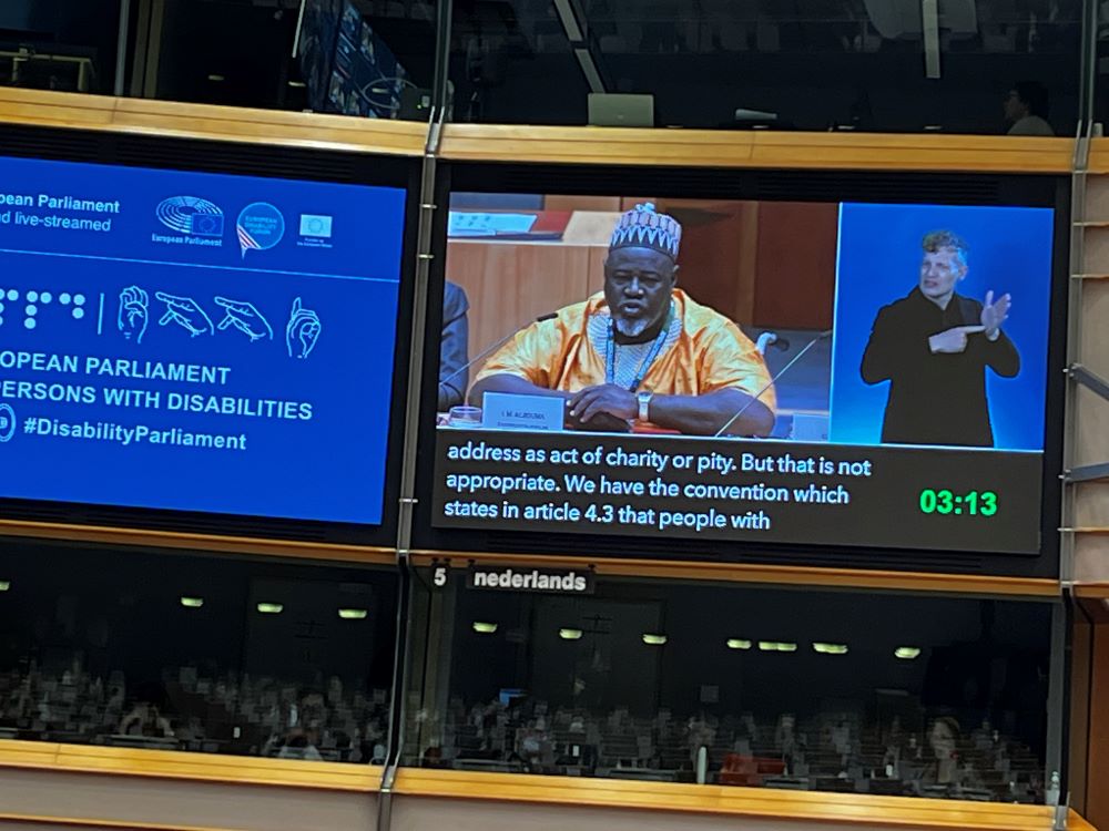 Predseda Afrického združenia osôb so ZP za rečníckym pultom. Na sebe má typický africký odev. Na fotografii je vidieť obrovský monitor, kde sa premieta vystúpenie. V spodnej časti vidieť prepis prejavu v reálnom čase.   