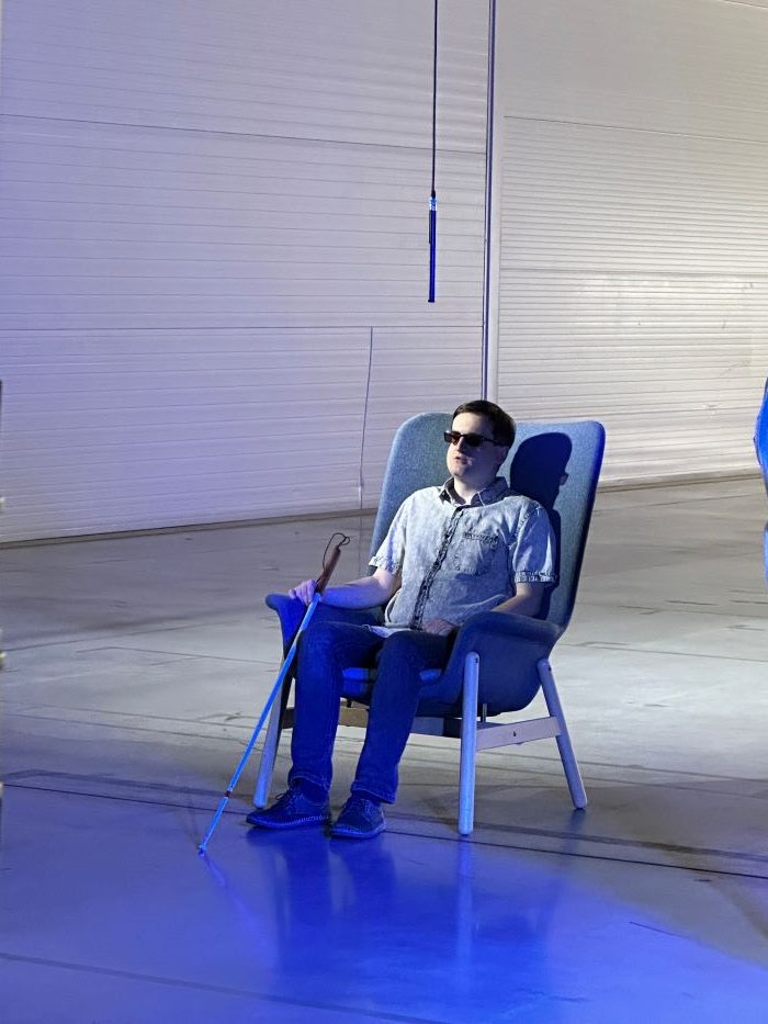 Na fotke je nevidiaci muž sediaci na stoličke s bielou palicou a modré efektné pozadie