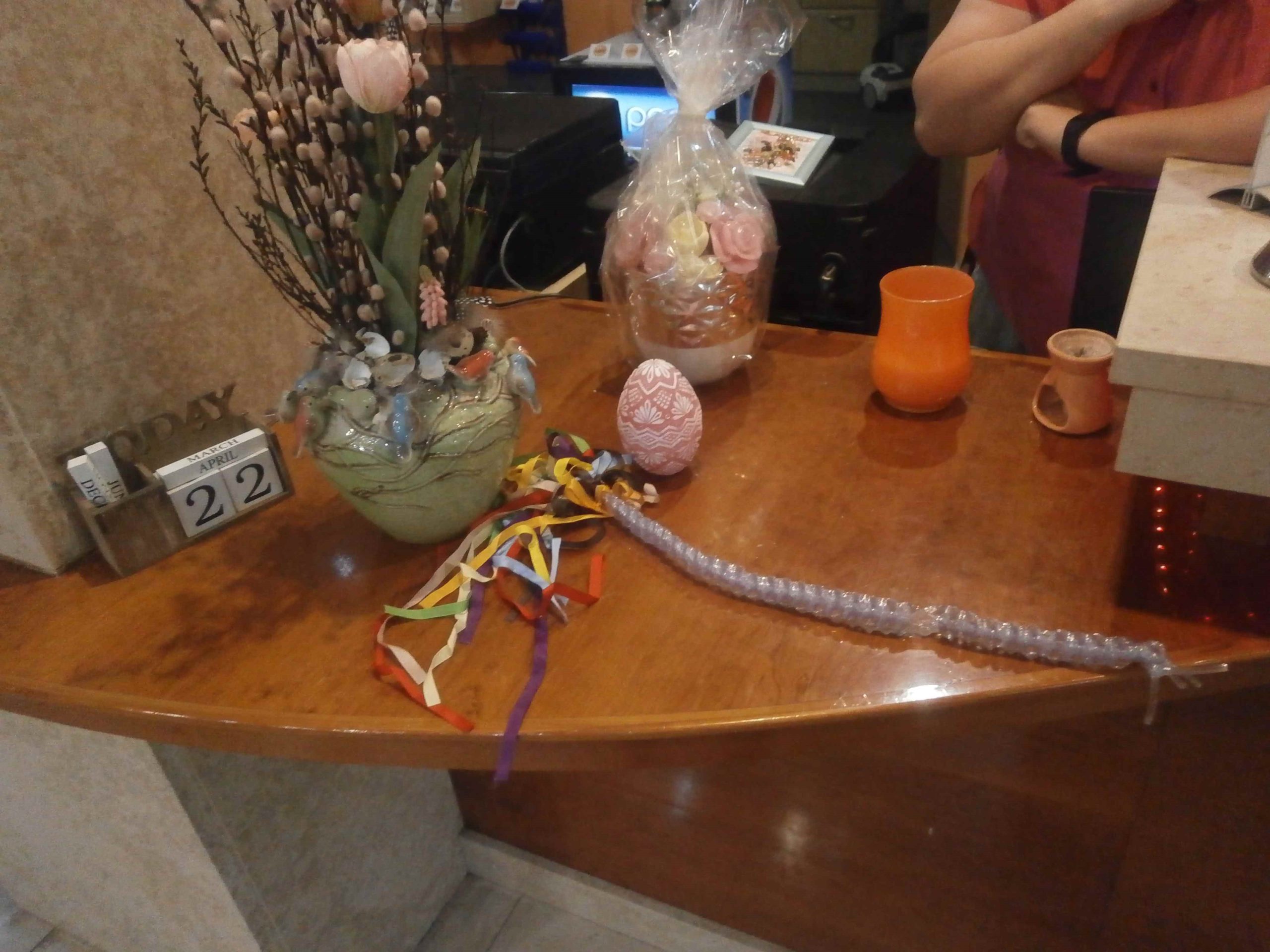 Stôl na ktorom je váza s veľkonočnou výzdobou, vedľa ktorej leží korbáč, ktorý je vyrobený s inhalačných hadičiek, zdobený farebnými stužkami.