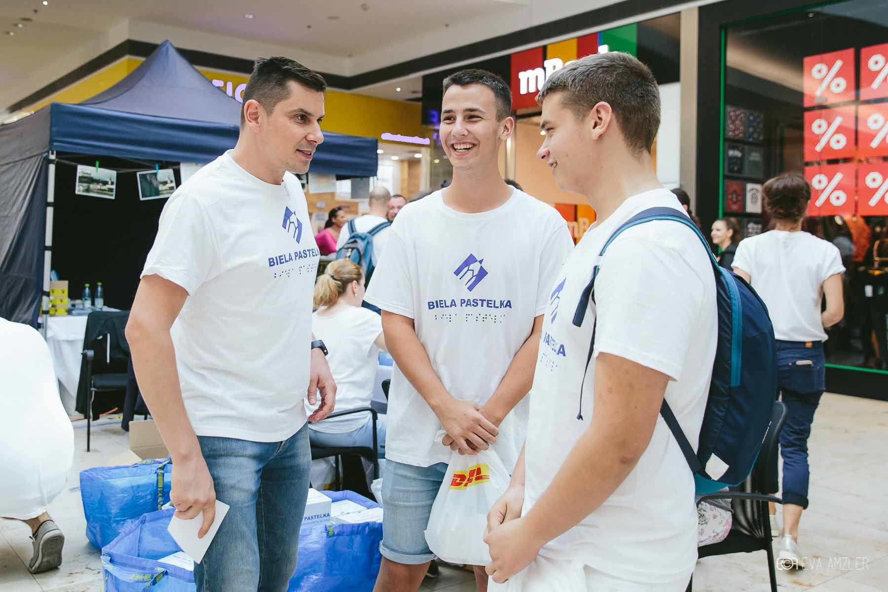 Dvaja muži dobrovoľníci v tričkách Biela pastelka v rozhovore s moderátorom Bielej pastelky.