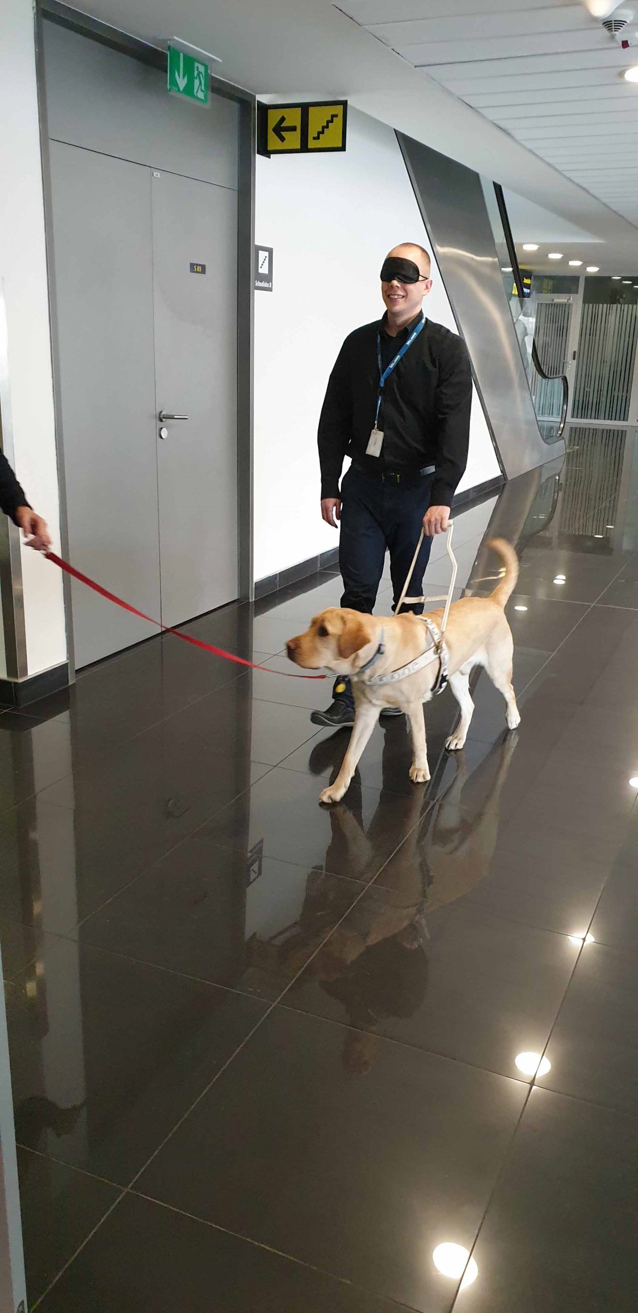 Muž s klapkami na očiach a vodiacim psom kráčajúci po letiskovej hale.