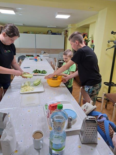 Tri deti v kuchyni. Jedno čistí zeleninu do plnky, ďalšie dve strúhajú.  