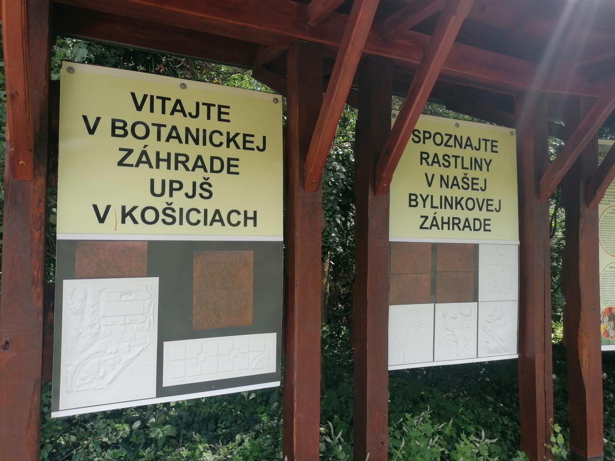 Na obrázku sú dve informačné tabule s nápismi Vitajte v Botanickej záhrade UPJŠ v Košiciach a Spoznajte rastliny v našej bylinkovej záhrade. Na tabuliach sú umiestnené aj reliéfne mapky a text v braili.  
