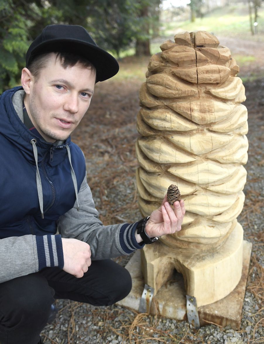 Marek Pizňak čupí pri veľkej drevenej plastike šišky. V ruke drží šišku v reálnej veľkosti.  