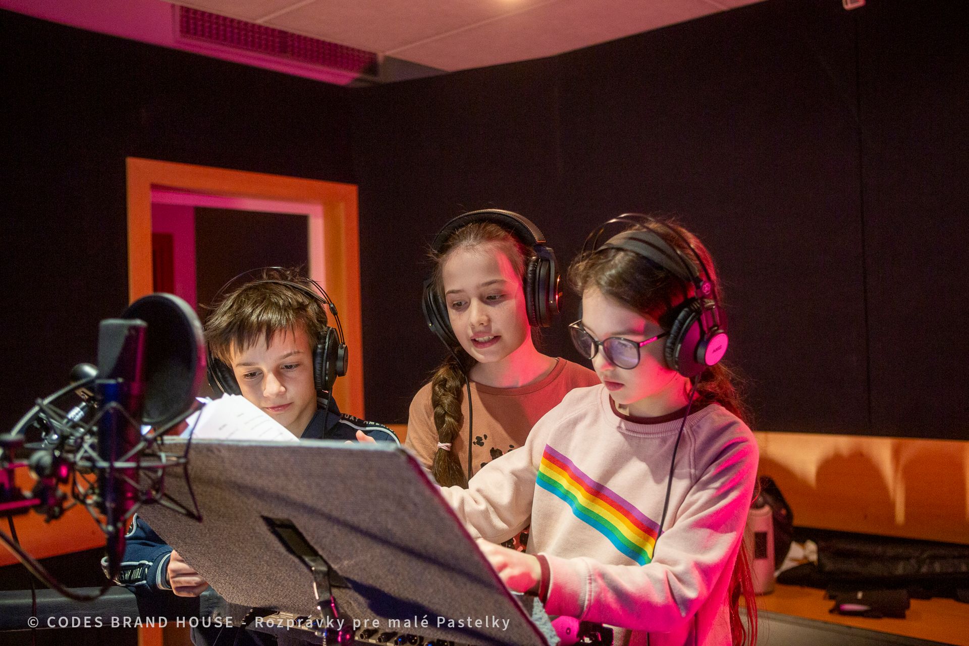 Tri deti (dve dievčatá a chlapec) sedia v štúdiu za mikrofónmi so slúchadlami na ušiach. Sú maximálne sústredení na prácu.
