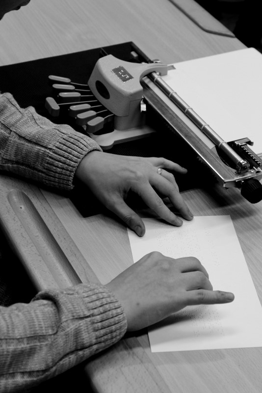 Ruky kontrolujúce text v braillovom písme. Vedľa je písací stroj pre braillovo písmo.