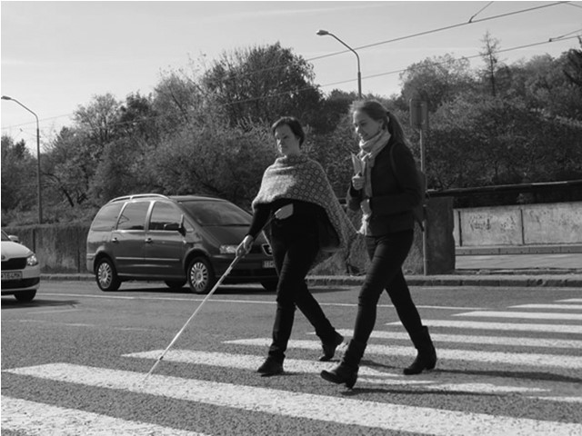 Nevidiaca žena s bielou palicou a so sprievodom prechádzajú cez prechod pre chodcov. 