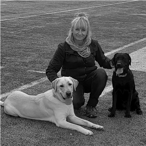 Žena na fotografii v strede pózuje s dvoma vodiacimi psami. Čierny na ľavo a biely napravo od nej. 