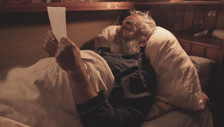 Scéna z filmu, na ktorej leží starší, šedivý, bradatý muž v posteli a číta z papiera. 