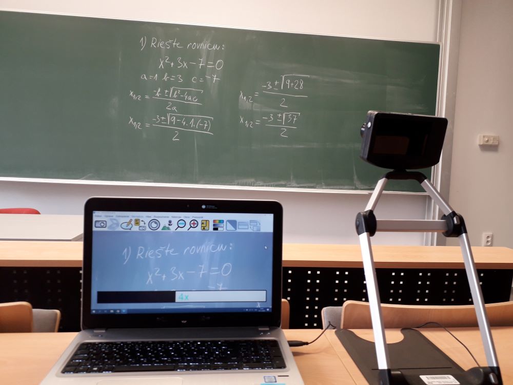 Na zábere vidíme obrazovku počítača, na nej sa zobrazuje školská tabuľa s časťou matematického zápisu. Za obrazovkou počítača je reálna tabuľa v učebni s celým zápisom. Vedľa počítača je na stole umiestnená kamera. 