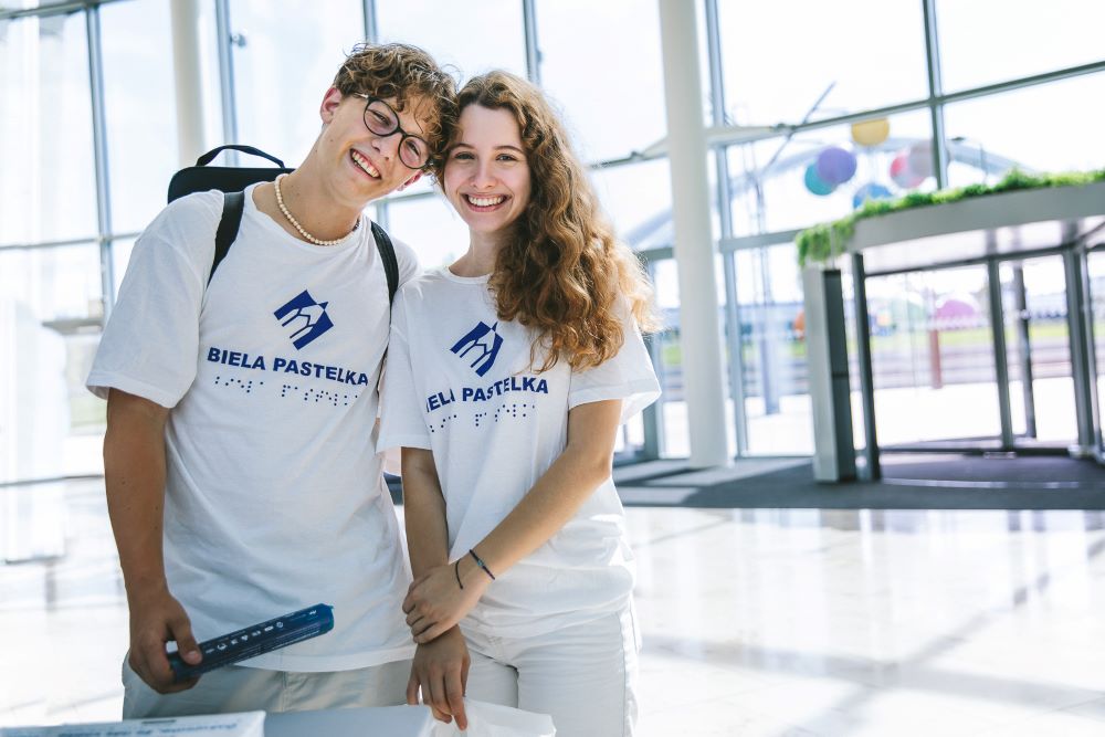 Na svetlom zaliatej fotografii stoja dvaja študenti s úsmevom na tvári a v tričkách s logom Bielej pastelky.  
