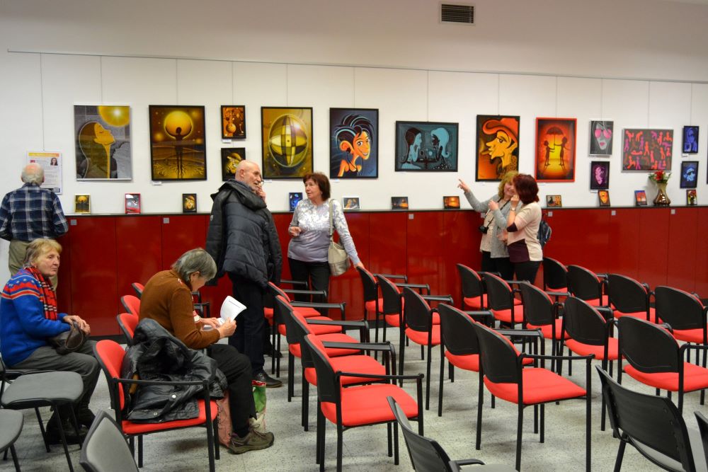 Záber z vernisáže – v miestnosti sú stoličky usporiadané do divadelného sedenia. Pár ľudí sa medzi sebou rozpráva. Na stenách visia obrazy.  
