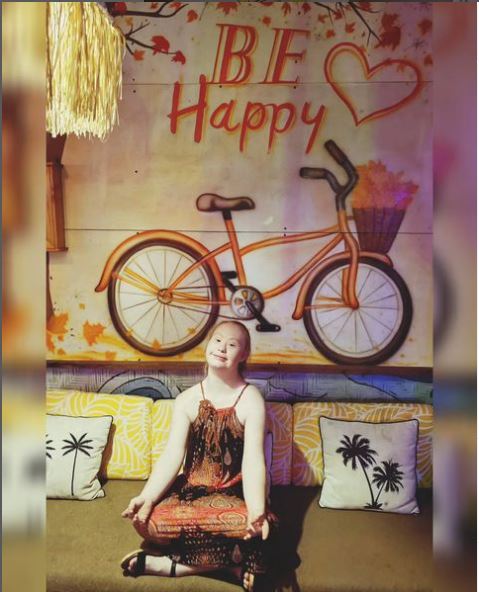 Mladá žena s Downovým syndrómom sedí v tureckom sede na gauči s vankúšmi. Na sebe má hnedé šaty na ramienka. Za ňou je obraz, na ktorom je bicykel a nápis Be happy a srdce. 
