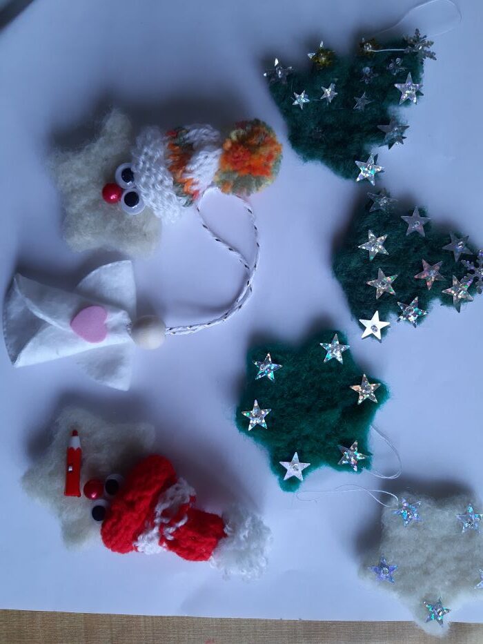 Plstené vianočné ozdoby - biele hviezdičky s tvárami a pletenými čiapkami, zelené hviezdičky a ihličnaté stromčeky ozdobené nalepenými striebornými hviezdičkami.  