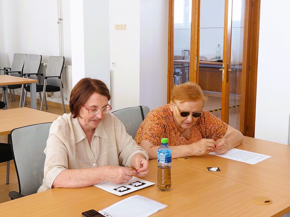 Pani Muščíková s ďalšou ženou sedia za stolom, pred nimi je rozložené hmatové pexeso. 