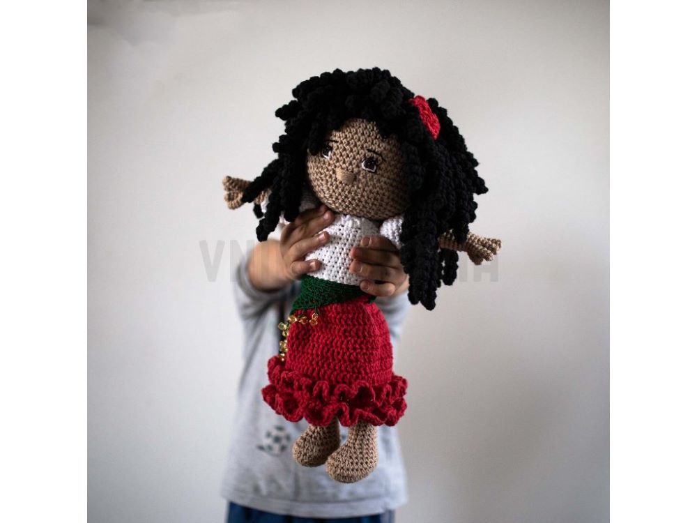 Na fotografii je háčkovaná bábika s dlhými čiernymi kučeravými vlasmi. Má tmavšiu pleť, oblečenú má bielu blúzku a dlhú červenú sukňu s volánom a šatkou previazanou v páse.