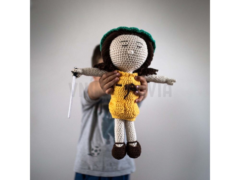 Na fotografii je háčkovaná bábika Tmejka. Oblečené má žlté šaty a zelenú baretku. Má zatvorené oči a v ruke drží bielu palicu.