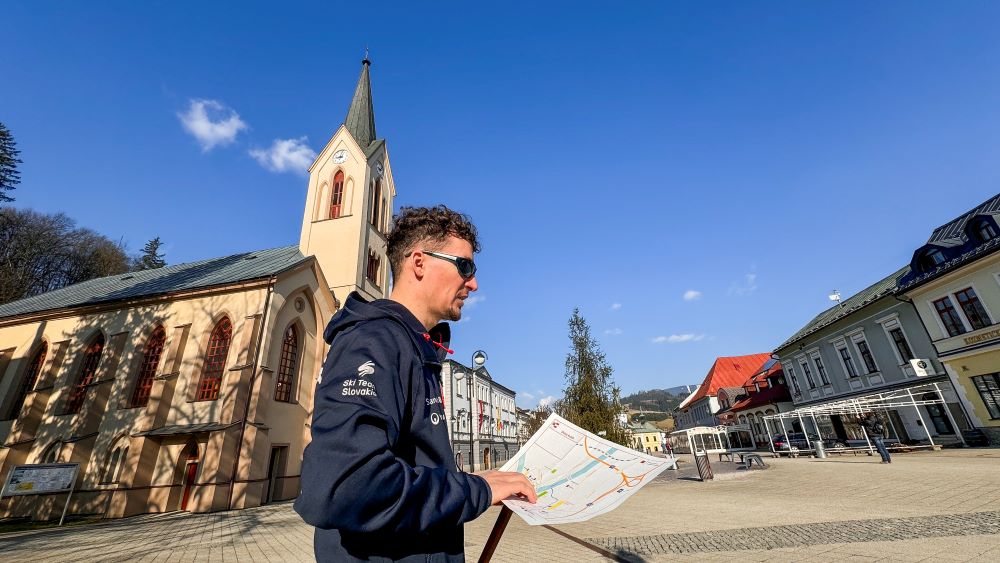 Na fotografii je nevidiaci mladý muž s reliéfnou mapou v ruke. Za ním vidieť kostol a domy na námestí.