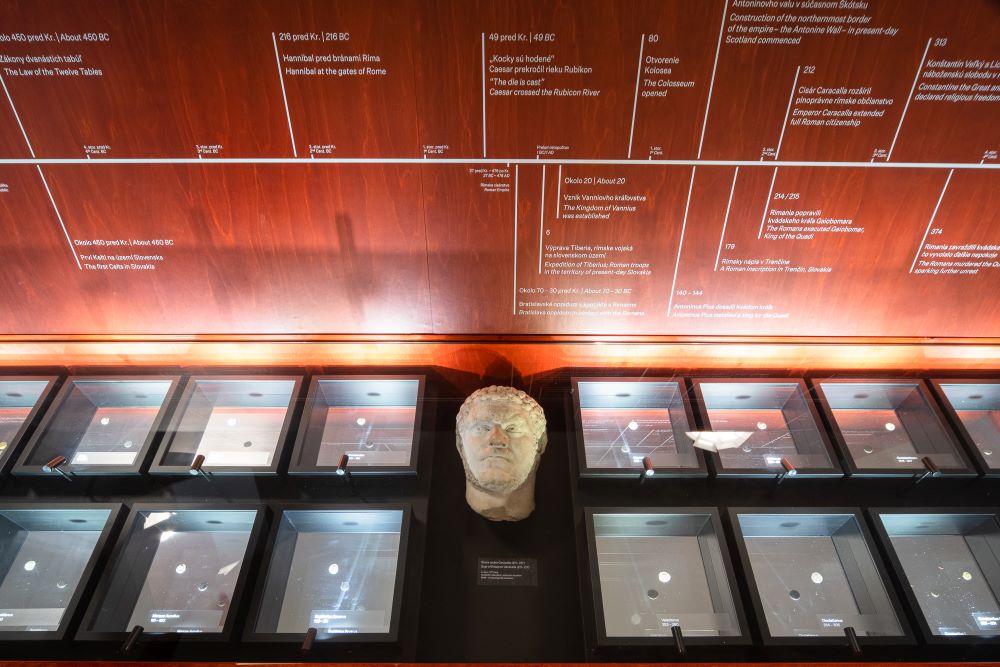Na fotografii sú nasvietené menšie vitríny s mincami, medzi nimi je busta cisára Caracallu. Na stene obloženej drevom za vitrínami je časová os. 