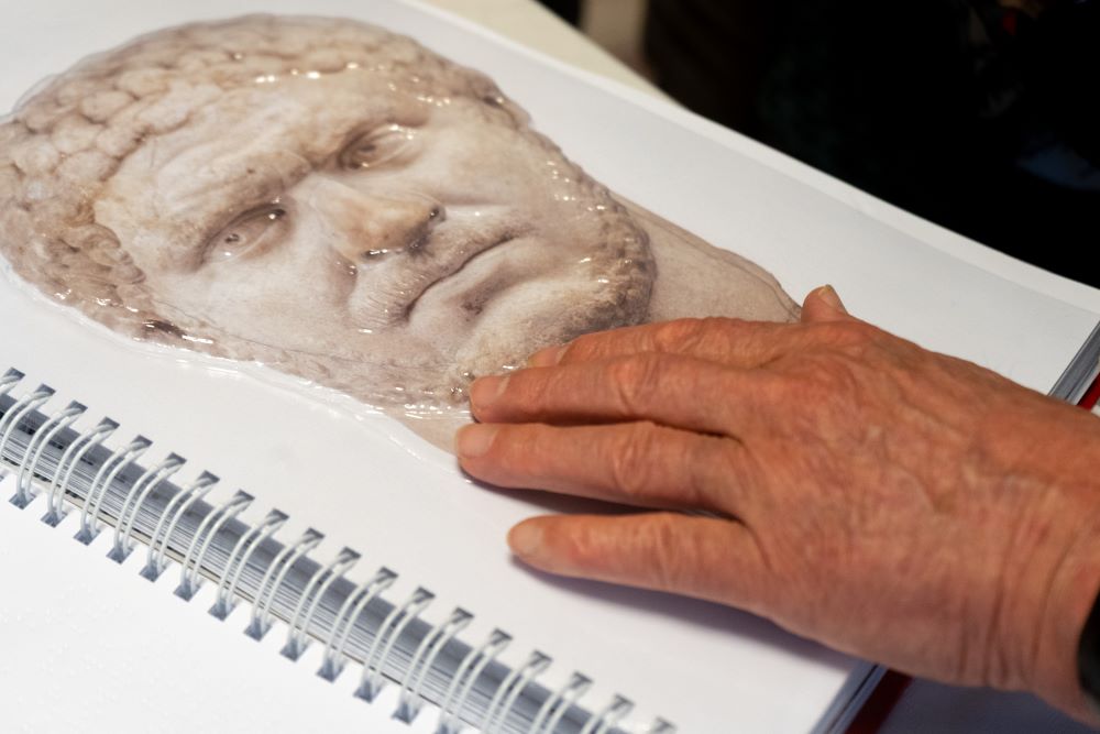 Záber na otvorenú knihu. Ruka sa dotýka reliéfnej podoby portrétu cisára Caracallu.