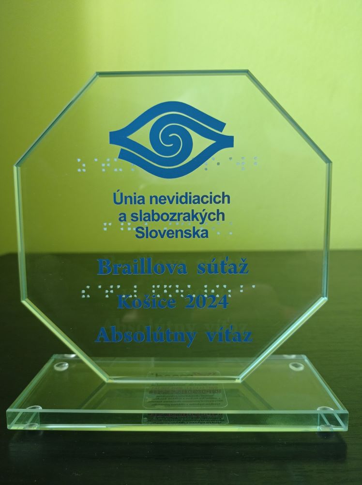 Detailný záber na sklenenú plaketu - na priehľadnom osemuholníku upevnenom na tenkom obdĺžnikovom podstavci je modré reliéfne logo ÚNSS, nápis Únia nevidiacich a slabozrakých Slovenska a text Braillova súťaž Košice, absolútny víťaz. Rovnaký text je na plakete aj Braillovým písmom.