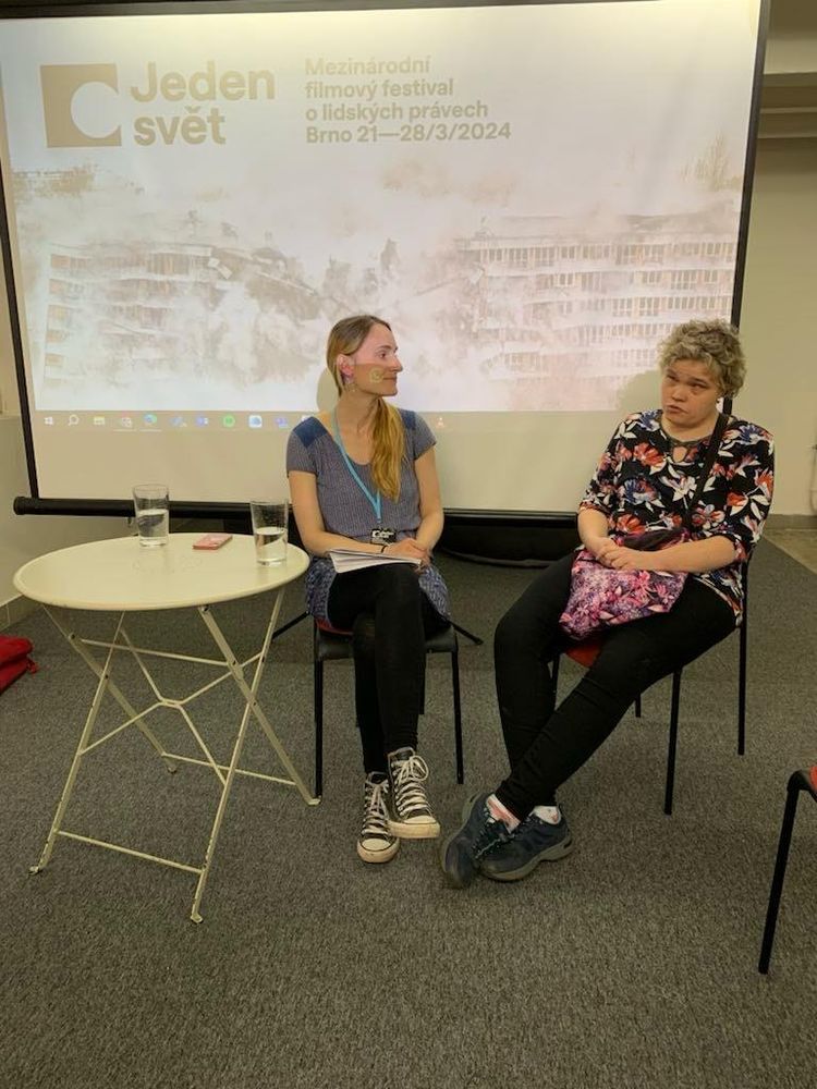 Pred plátnom s prezentáciou sedí Lucia a moderátorka. Pred nimi je jednoduchý stolík s pohármi s vodou. 