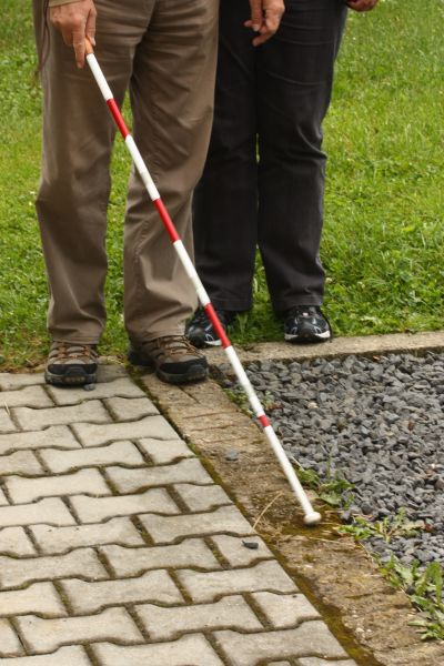 Na fotografii vidíme nohy dvoch osôb. Jedna z nich drží červeno-bielu palicu a prezerá si ňou obrubník pred sebou. 