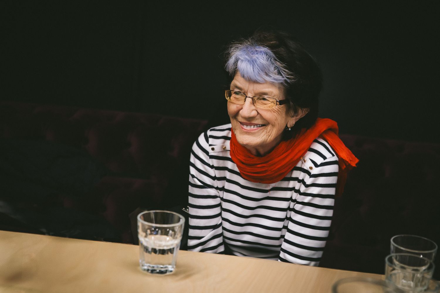 Za stolom sedí usmievavá staršia žena v prúžkovanom tričku s červenou šatkou okolo krku. Má krátke tmavé vlasy s fialovou ofinou. Pred ňou na stole je položený pohár vody. 