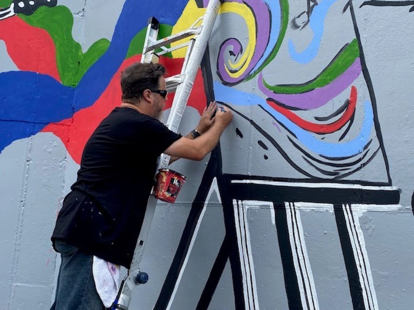 Muž v čiernom tričku stojí na rebríku opretom o múr. K rebríku je pripevnená plechovka s farbou, muž drží v ruke štetec. Na múre je rozmaľovaný obraz, muž na ňom práve pracuje. Obraz je abstraktný a veľmi farebný. 