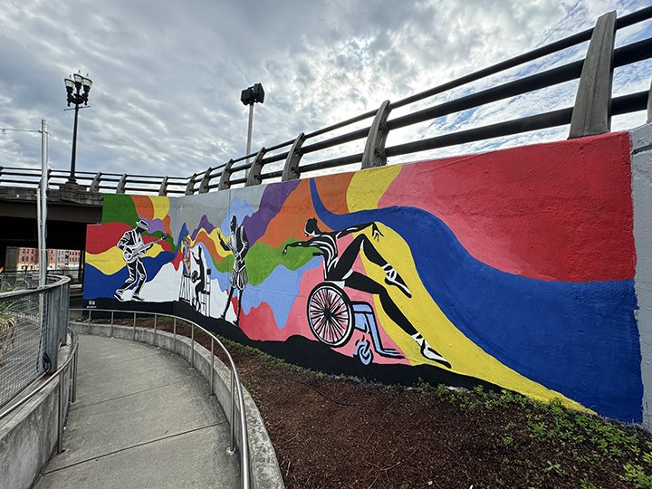 Záber na veľkú nástennú výrazne farebnú maľbu. Na nej sa dá rozoznať niekoľko postáv – žena v baletných špičkách a v tanečnej póze na invalidnom vozíku, muž s gitarou v ruke, či maliar sediaci pred plátnom na stojane. Pozadie obrazu tvoria abstraktné farebné plochy.