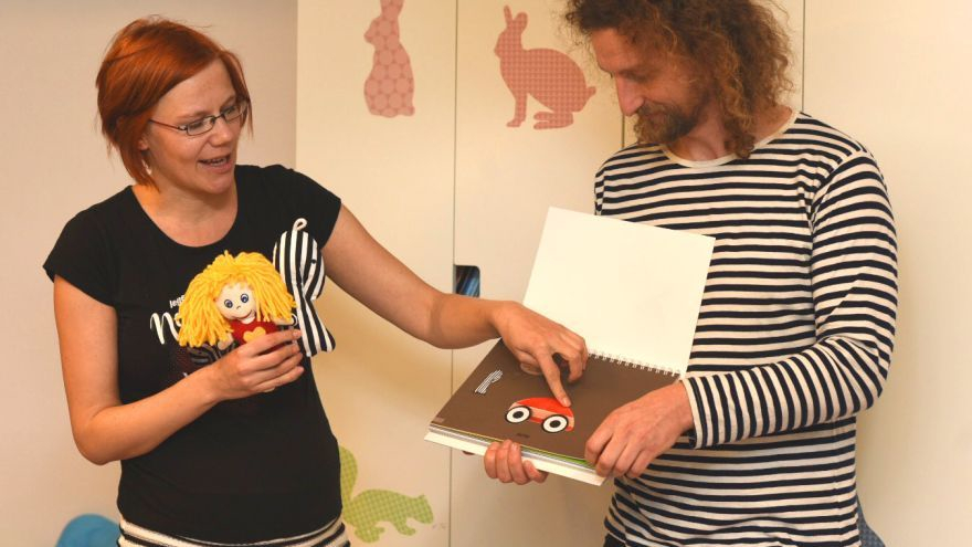 Na fotografii je mladšia žena s ryšavými vlasmi v čiernom tričku a čierno-bielej prúžkovanej sukni s bábikou a prúžkovanou bábkou v ruke. Muž vedľa nej má dlhšie kučeravé vlasy a bradu. Oblečené má prúžkované tričko. V ruke drží knihu otvorenú na strane s reliéfnym obrázkom autíčka. Žena si rukou prezerá autíčko. 