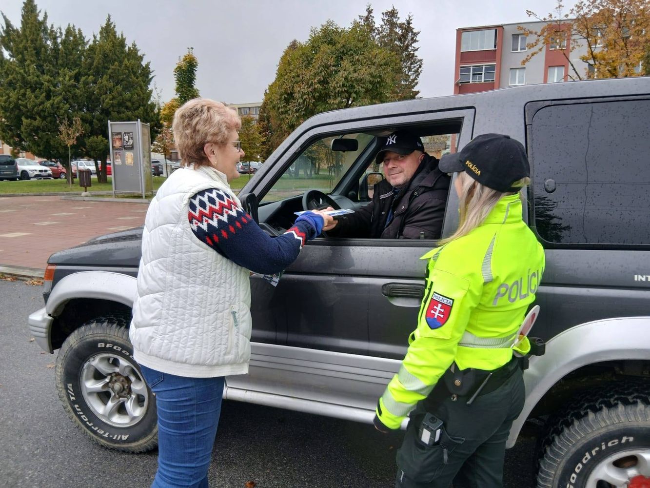 Začiatok alternatívneho popisu. Na ceste stojí auto so stiahnutým oknom, za volantom sedí vodič. Vedľa auta stojí policajtka a staršia žena, ktorá vodičovi niečo podáva. Koniec alternatívneho popisu.