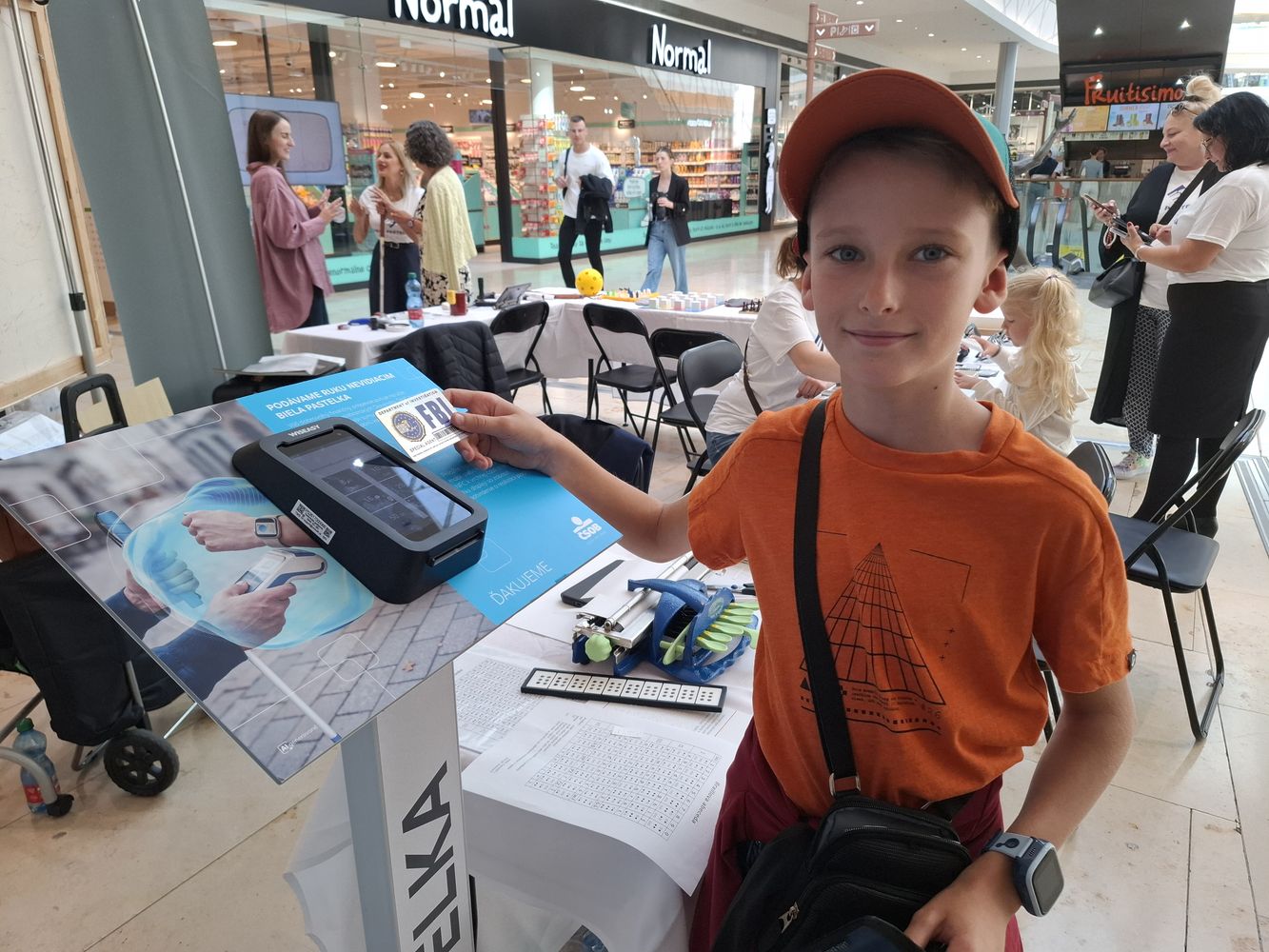 Začiatok alternatívneho popisu. Na fotografii je asi 10-ročný chlapec v šiltovke. Stojí pri termináli a prikladá k nemu platobnú kartu, ktorá vyzerá ako služobná karta agenta FBI. Terminál je umiestnený na stojane s nápisom Biela pastelka medzi infostánkami ÚNSS v nákupnom centre. Koniec alternatívneho popisu.